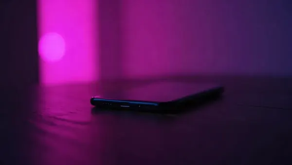 Celular sobre uma mesa de madeira escura, cercado de reflexos violeta e rosa, ilustração do artigo “Polybuzz em português 2026: grátis, regras e alternativas”